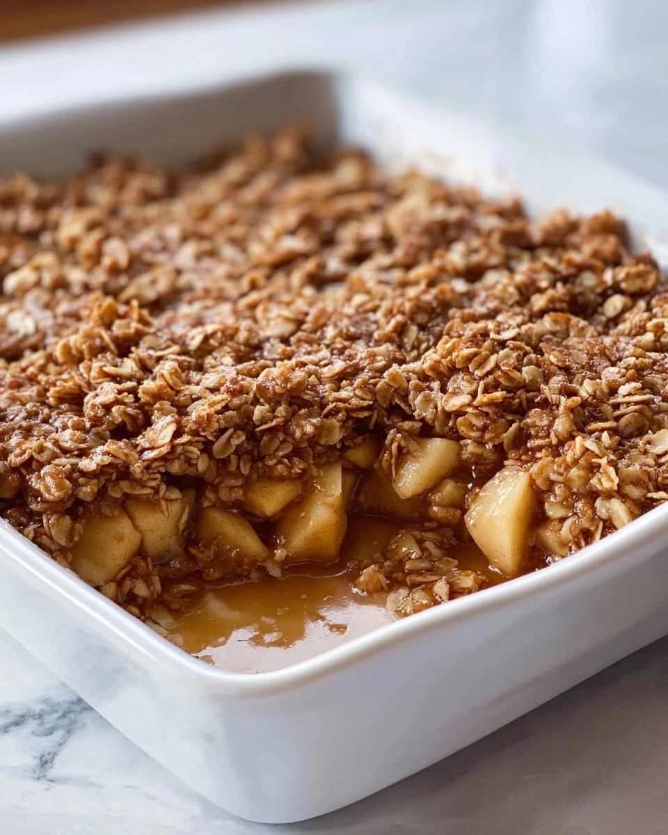 The image shows a baked apple crisp in a white square baking dish, placed on a white marbled surface. The dish has two main layers: the bottom layer consists of chunky apple pieces mixed with oats and a syrupy brown sauce, creating a soft and juicy texture; on top, there is a thick, crumbly oat and spice topping with a golden brown color, giving a crunchy look. The edges of the dish show the layers clearly, and the topping has a slightly glossy finish from the syrup. Photo taken with an iphone --ar 4:5 --v 7