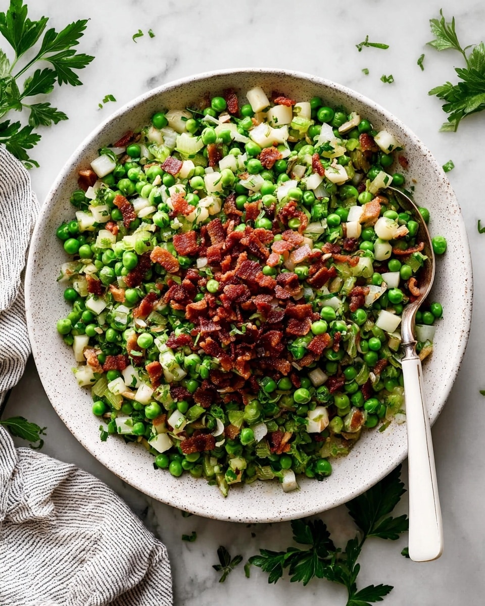 A white speckled plate is filled with a vibrant salad that shows different layers and colors. The bottom layer is made of bright green peas scattered all over, mixed with small pale yellow potato cubes and thin strips of translucent cooked onions, garnished with bits of fresh green parsley. On top, there is a thick mound of small, crispy, reddish-brown bacon pieces placed in the center, adding a strong warm color contrast. A shiny silver spoon with a white handle rests on the plate, slightly embedded in the salad from the right side. The plate is set on a white marbled textured surface with some scattered pepper and parsley around, and a striped cloth napkin is partly visible on the bottom right. photo taken with an iphone --ar 4:5 --v 7