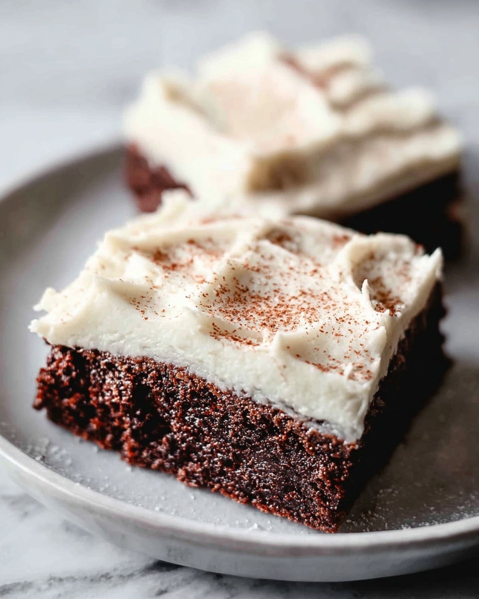 The image shows two square pieces of chocolate cake with a thick layer of creamy white frosting on top, sitting close to each other on a white plate. The chocolate cake layer is dark brown with a moist and slightly crumbly texture. The frosting on top is smooth with swirls and small ridges, and it is lightly sprinkled with a fine dusting of brown powder. The plate holding the cakes has a simple design and rests on a white marbled surface. photo taken with an iphone --ar 4:5 --v 7
