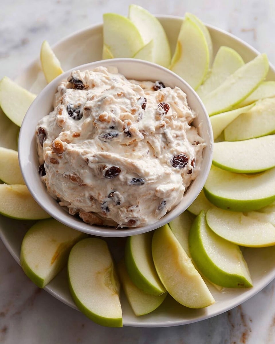 A white bowl filled with creamy, light tan dip that has visible dark raisins and small nut pieces mixed throughout, showing a thick and textured consistency with soft peaks on the surface. The bowl is placed on a white plate, which is surrounded by fresh green apple slices showing white flesh and smooth green skin, arranged loosely around the bowl. The setting features a white marbled surface in the background. photo taken with an iphone --ar 4:5 --v 7