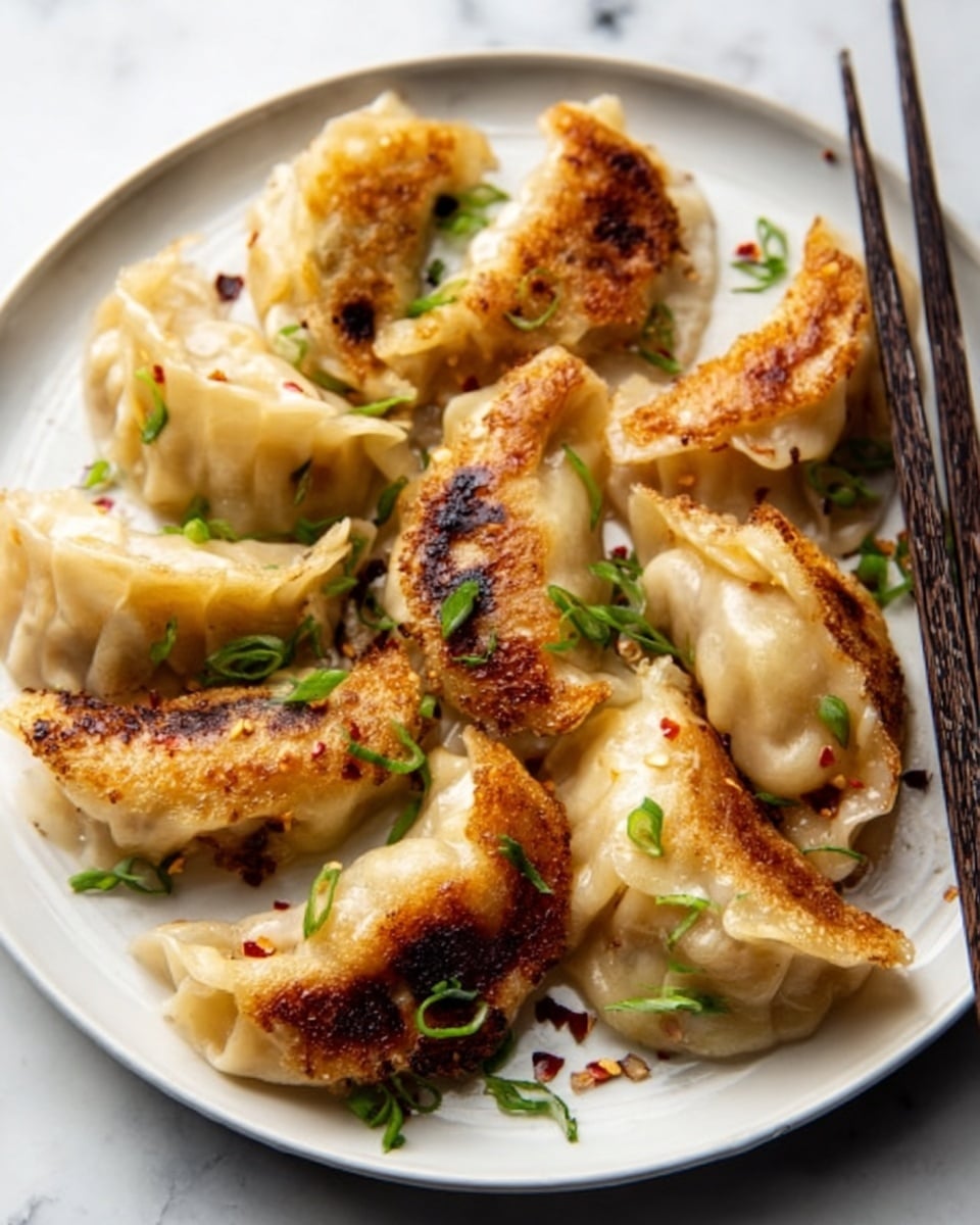 A white plate holds a single layer of golden brown pan-fried dumplings arranged in a loose circle. The dumplings have slightly crispy edges with some darker browned spots and soft, slightly translucent dough on top. They are sprinkled with small green chopped scallions and red chili flakes. A pair of dark brown chopsticks rests diagonally on the plate, and the plate sits on a white marbled surface. photo taken with an iphone --ar 4:5 --v 7
