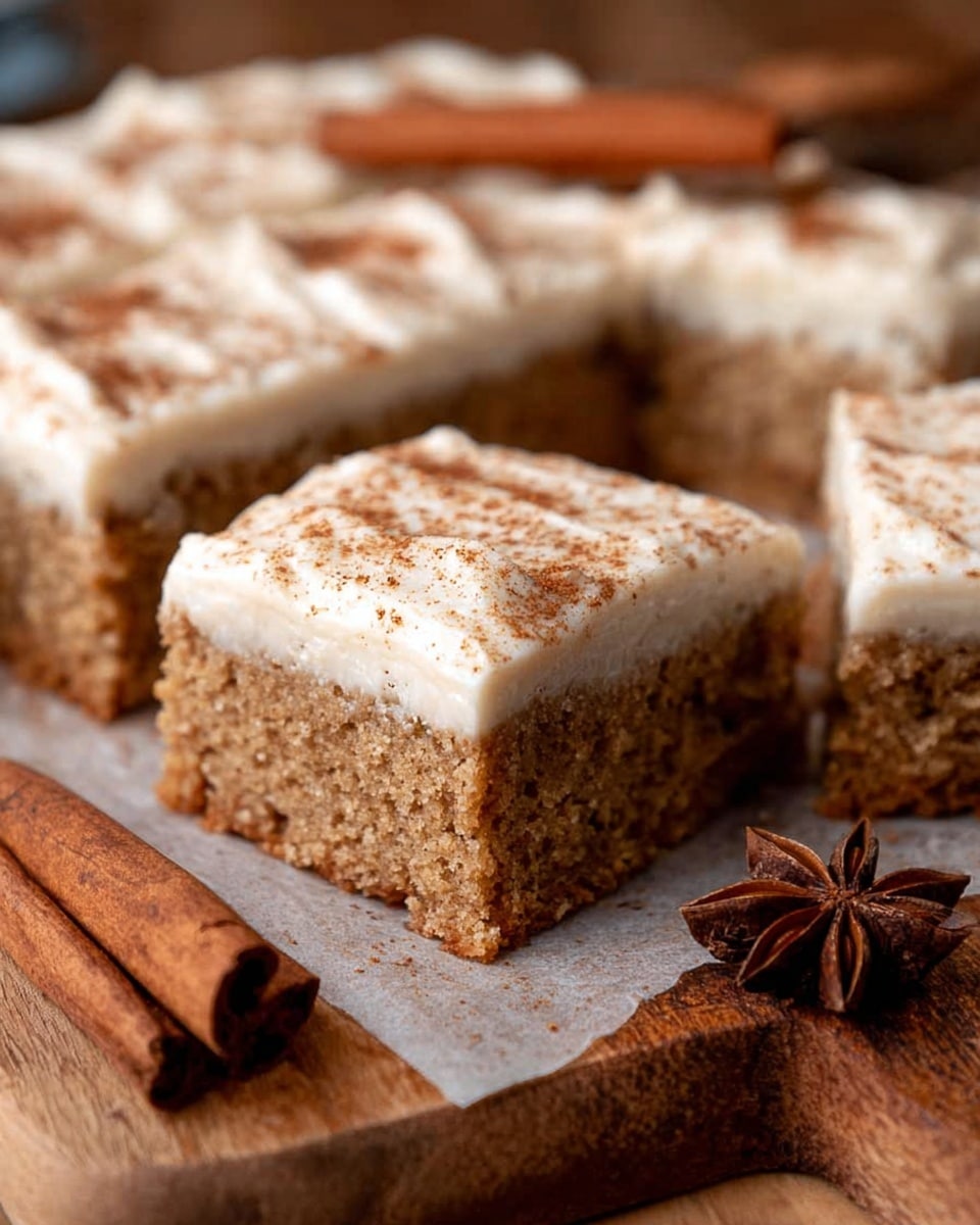 A close-up image of square cake pieces with two distinct layers: the bottom layer is a thick, light brown, moist sponge cake with a soft texture, while the top layer is a creamy, light beige frosting sprinkled lightly with brown spices on top. The cake squares are placed close together on white parchment paper over a wooden surface, with visible cinnamon sticks and star anise around them, all set against a white marbled textured background. photo taken with an iphone --ar 4:5 --v 7