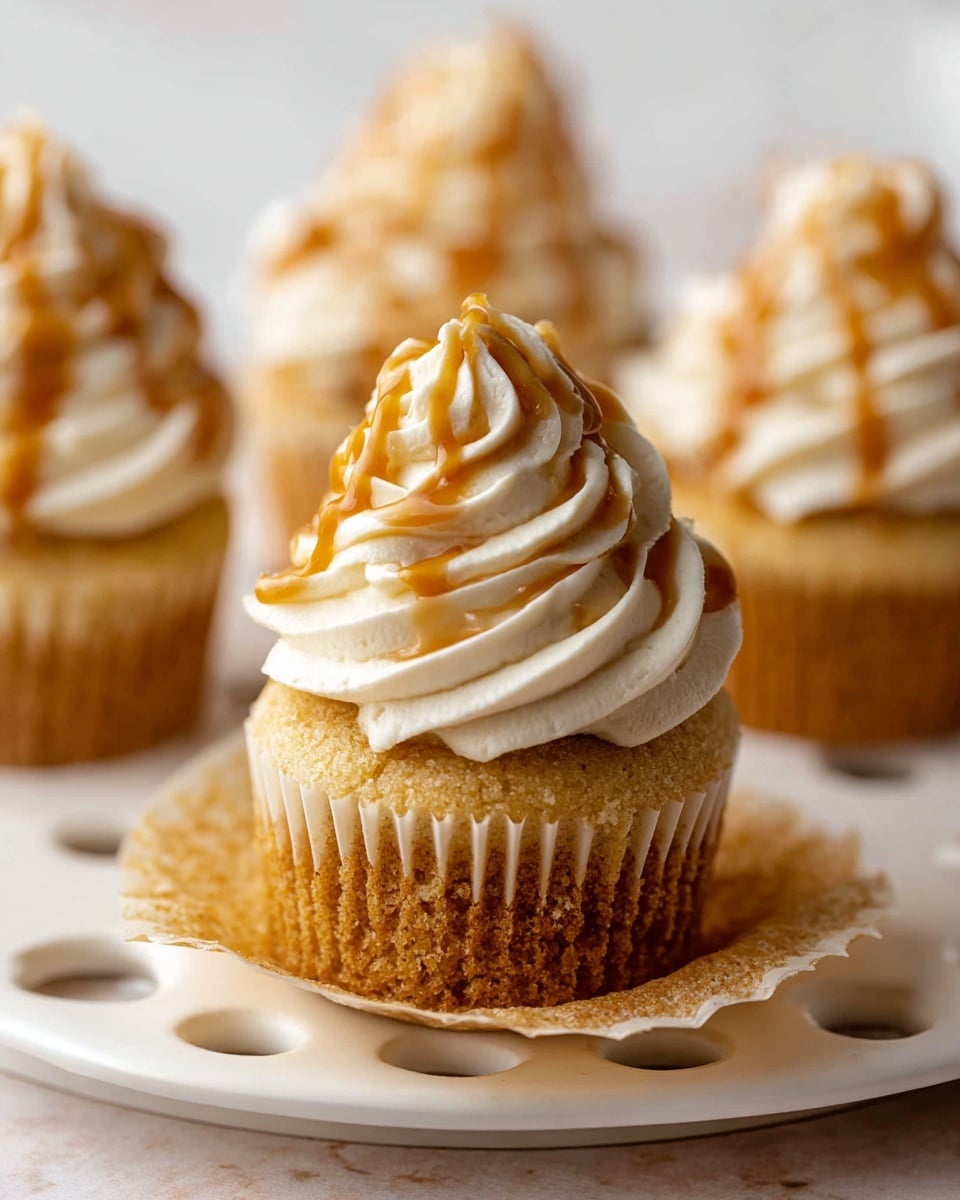 A close-up view of a cupcake with two main layers: a golden brown cake base with a soft, slightly crumbly texture sitting on an open beige paper liner, topped by a tall swirl of creamy off-white frosting with a smooth and fluffy texture, drizzled with a glossy light brown caramel sauce that lightly runs down the frosting’s curves. In the background, three more identical cupcakes are slightly out of focus, also topped with swirled frosting and caramel drizzle, all placed on a white plate with round indentations along the edge, all set on a white marbled surface. photo taken with an iphone --ar 4:5 --v 7
