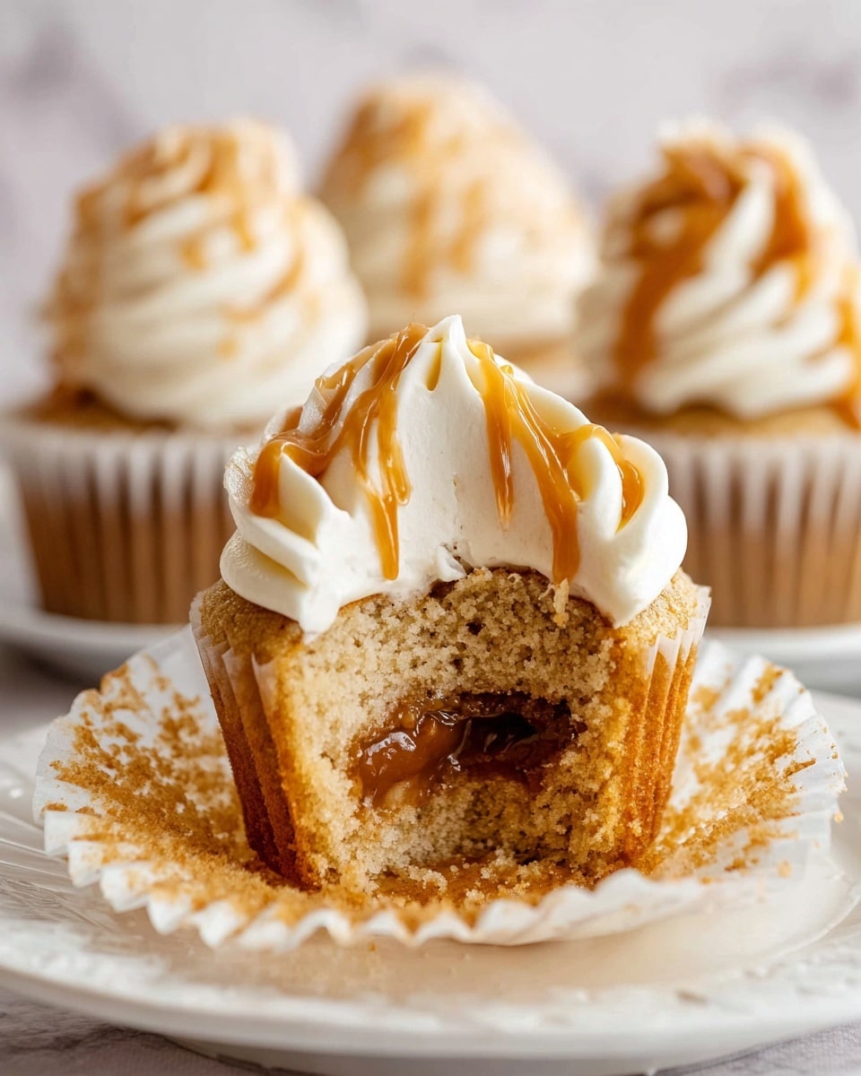 A close-up of a cupcake that has been cut in half showing three layers: the bottom layer is a moist light brown cake, the middle layer is a gooey caramel filling with a smooth texture, and the top layer is a thick swirl of creamy white frosting with caramel drizzle on the side. The cupcake is resting on a white cupcake liner that is open and sits on a white plate with a delicate scalloped edge, all placed on a white marbled surface. In the blurred background, there are three whole cupcakes topped with the same white frosting and caramel drizzle. photo taken with an iphone --ar 4:5 --v 7