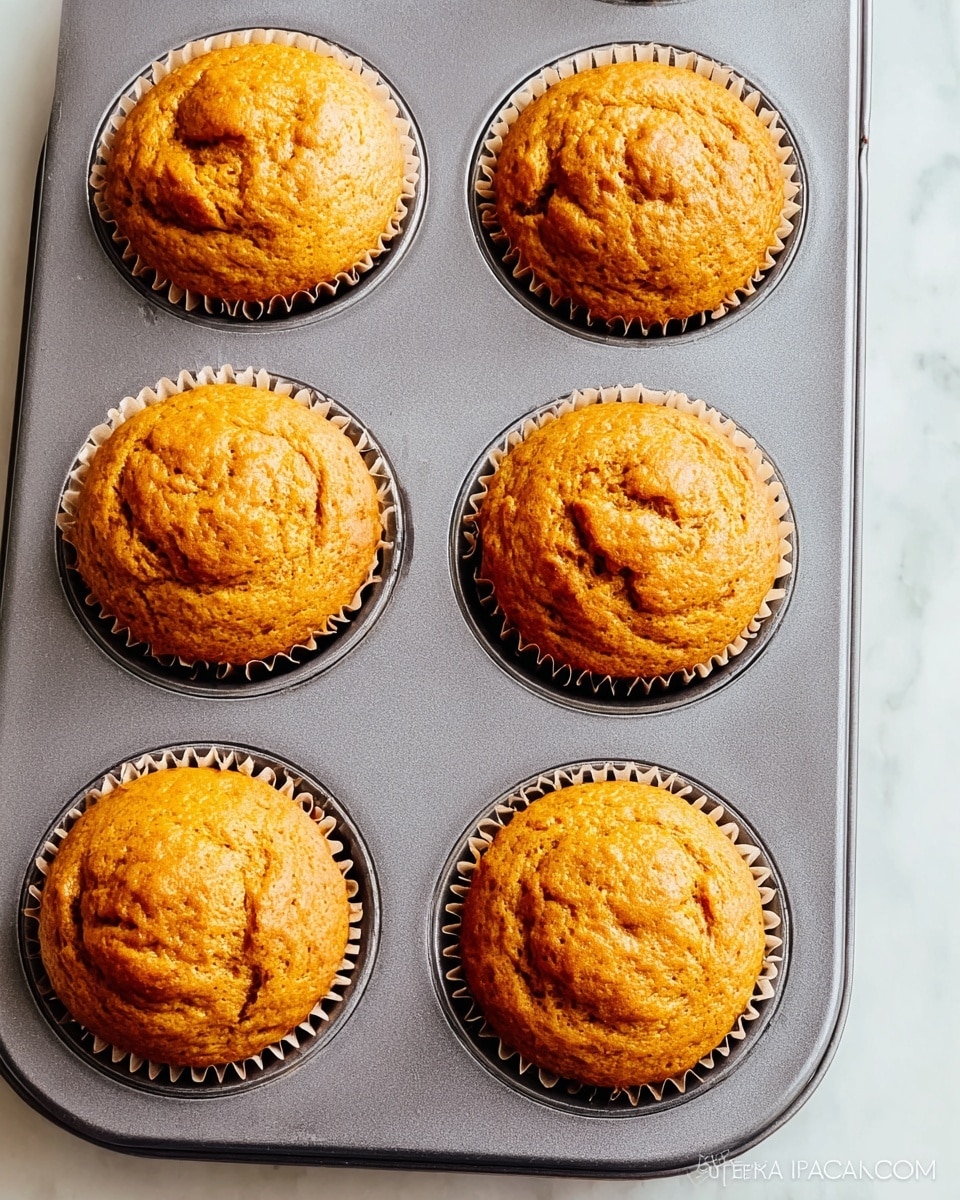 A close-up view of six golden-brown muffins baked evenly in a grey muffin tray, each muffin sitting in a light brown paper liner with slightly ruffled edges. The muffins are rounded on top with a textured, slightly cracked surface showing their soft, moist interior. The tray rests on a white marbled textured surface, highlighting the warm tones of the muffins. Photo taken with an iphone --ar 4:5 --v 7