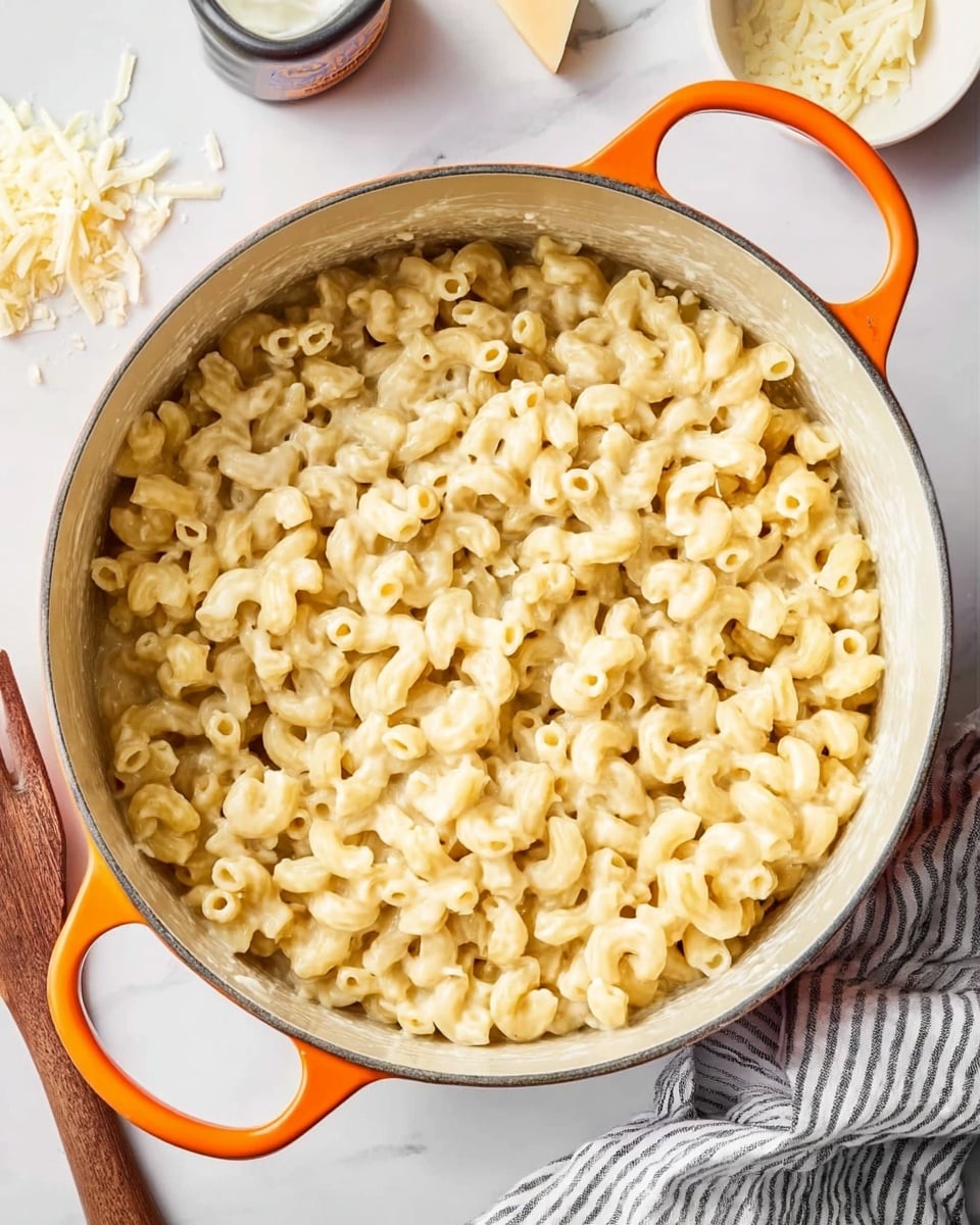 A large white pot with orange handles sits on a white marbled surface, filled to the top with creamy macaroni and cheese. The pasta is elbow-shaped, coated evenly with a smooth, pale yellow cheese sauce that looks rich and thick. A few small bits of cheese are scattered inside the pot, and some uncooked pasta pieces and white cheese shreds lie next to it on the surface. A white bowl and striped cloth napkin are placed nearby, adding a cozy touch to the scene. photo taken with an iphone --ar 4:5 --v 7