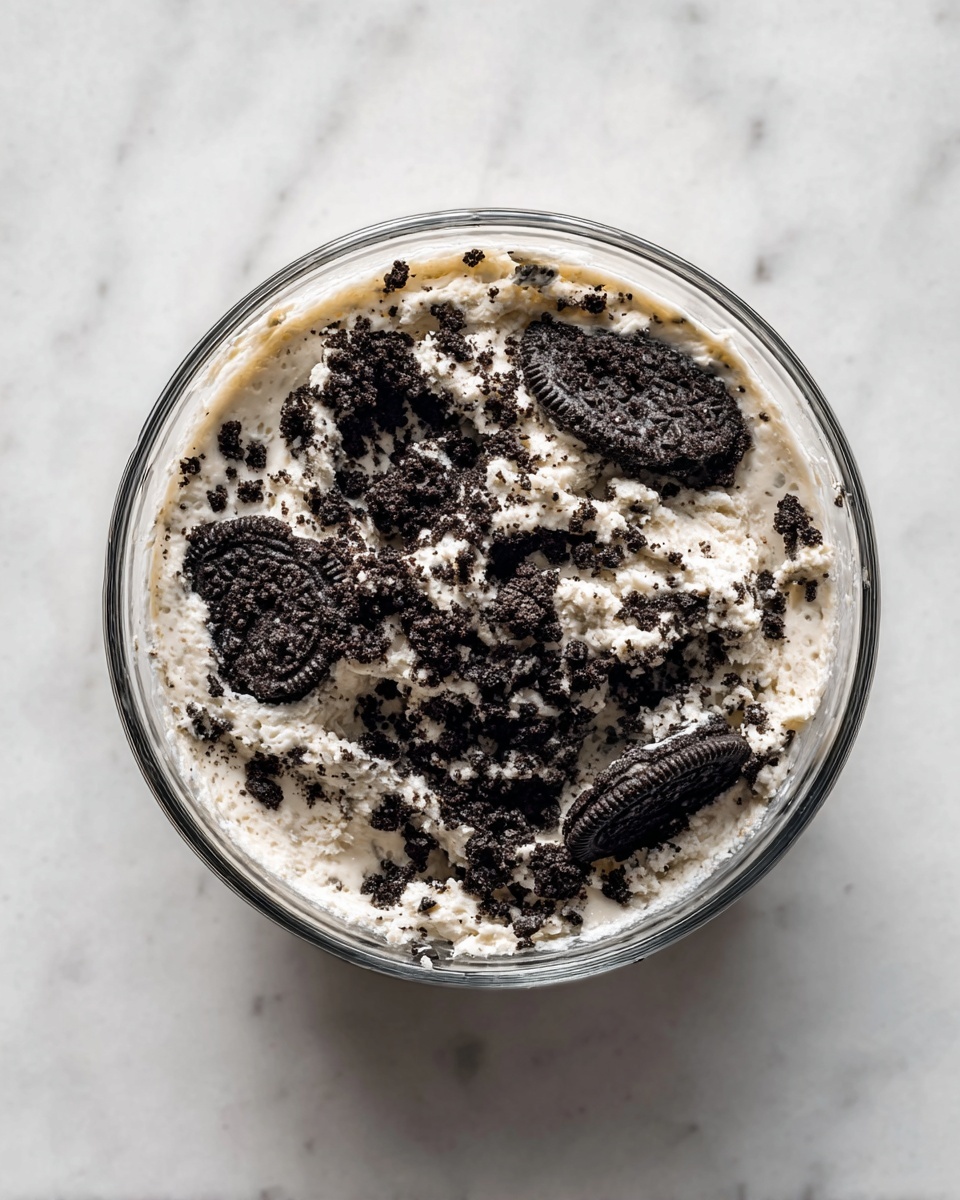A clear glass bowl filled with a mix of creamy white batter and large chunks of dark, crumbly chocolate cookie pieces. The batter has a smooth, slightly thick texture, with the black cookie bits scattered unevenly throughout, creating a speckled pattern. The bowl rests on a white marbled surface, highlighting the contrast between the light batter and dark cookie crumbs. photo taken with an iphone --ar 4:5 --v 7