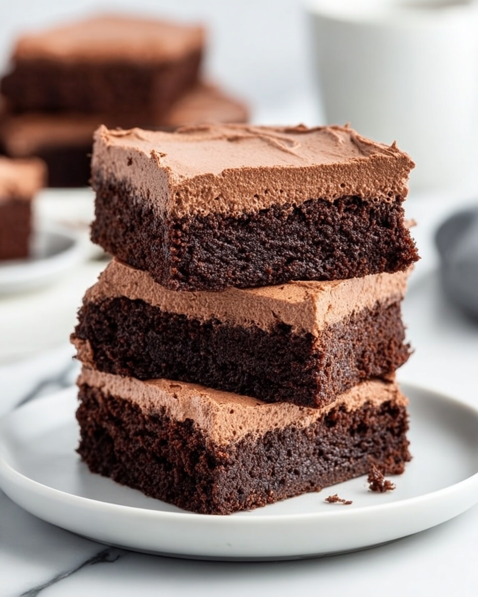 The image shows three square chocolate brownies stacked on a white plate, placed on a white marbled surface. Each brownie has two layers: a thick, dark brown, moist base and a lighter, fluffy chocolate frosting on top. The layers are even and smooth, with the frosting spread neatly across the surface. The brownies look rich and soft, with the edges of the base slightly crumbly. In the background, more brownies and a white cup are blurred out. Photo taken with an iphone --ar 4:5 --v 7