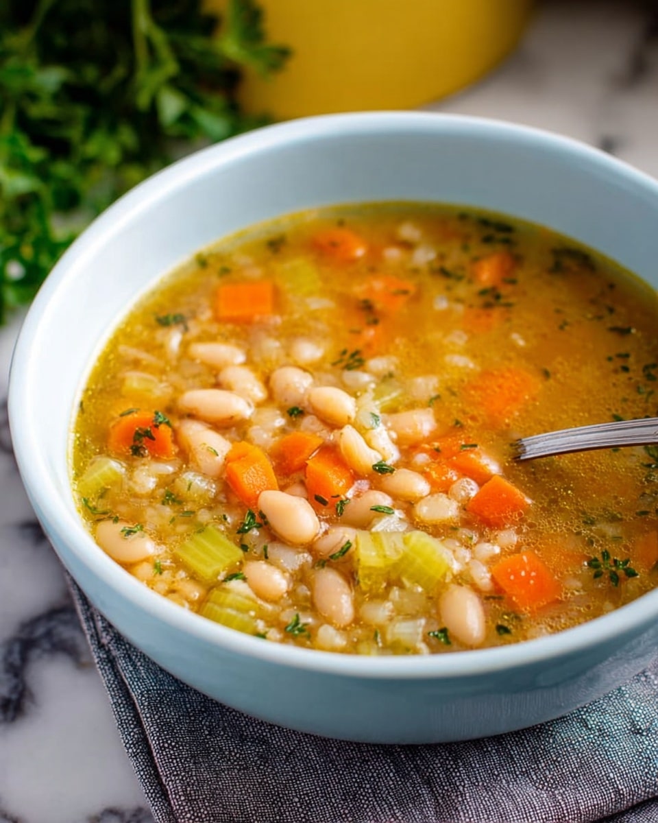 A bright blue bowl filled with a thick, chunky soup made up of three visible layers: the bottom layer is a clear golden broth with floating herbs, the middle layer contains pale white beans that are soft and round, and the top layer has diced orange carrots and green celery pieces scattered evenly across, with a silver spoon resting inside the bowl. The bowl is sitting on a gray cloth, all placed on a white marbled textured surface. Photo taken with an iphone --ar 4:5 --v 7