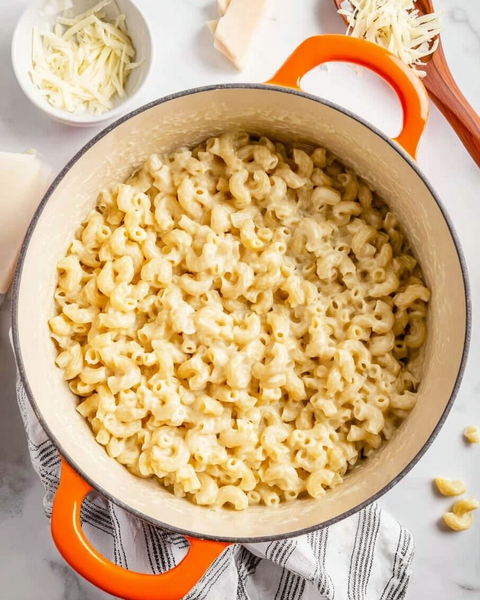 The image shows a white pot with orange handles filled with creamy macaroni and cheese. The macaroni noodles are small and curved, covered in a thick, smooth, pale yellow cheese sauce. The pot sits on a white marbled surface with a striped cloth next to it and some uncooked pasta pieces scattered nearby. Parts of white bowls are visible around the pot. The overall look is warm and inviting, focusing on the rich texture of the cheesy pasta. photo taken with an iphone --ar 4:5 --v 7