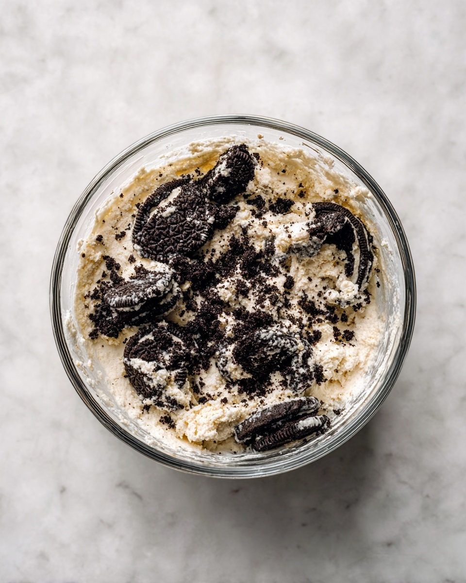 A clear glass bowl sits on a white marbled surface, filled with a mixture of crumbled black cookie pieces and a creamy white base. The texture is grainy and slightly chunky with generous amounts of dark cookie crumbs scattered unevenly throughout the creamy layer. Some larger chunks of cookies are visible, embedded within and resting on top of the mixture, giving a rough and varied appearance. The bowl's transparent sides reveal the mix densely packed inside, with traces of the creamy base stuck to the inner edges. photo taken with an iphone --ar 4:5 --v 7