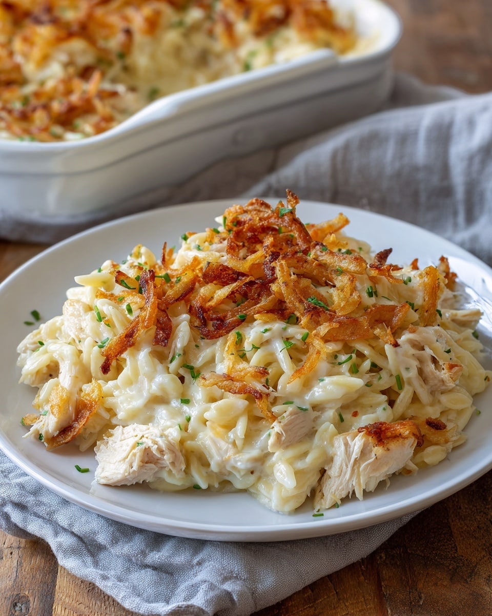 A white plate holds a creamy casserole dish with three visible layers: at the bottom, soft cooked orzo pasta mixed with tender chicken chunks in a thick, smooth white sauce; the middle layer maintains the creamy texture with bits of chicken and pasta blending throughout; on top, a generous layer of crispy, golden-brown fried onions scattered unevenly, adding contrast in color and texture, sprinkled with small bright green herb pieces for garnish. The plate is set on a white marbled surface with a soft gray cloth partially shown beside it, while a silver fork rests beside the food on the plate. In the blurred background, a large rectangular white baking dish filled with more of the casserole is visible. Photo taken with an iphone --ar 4:5 --v 7