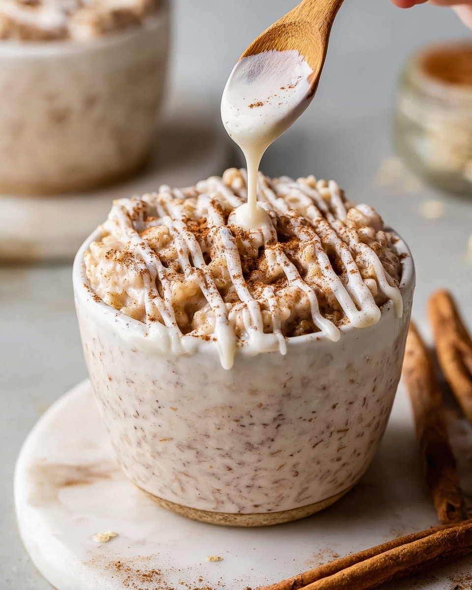 A small white cup filled with a soft, creamy rice pudding topped with a thick drizzle of white icing and a sprinkle of brown cinnamon powder. The rice pudding has a slightly textured, grainy look with visible rice grains. The icing creates thin, white lines over the surface, while the cinnamon adds a dusted, warm brown layer on top. A woman's hand holds a wooden spoon above the cup, dripping some pudding back into it. The cup sits on a white marbled surface with another similar cup blurred in the background. Photo taken with an iphone --ar 4:5 --v 7