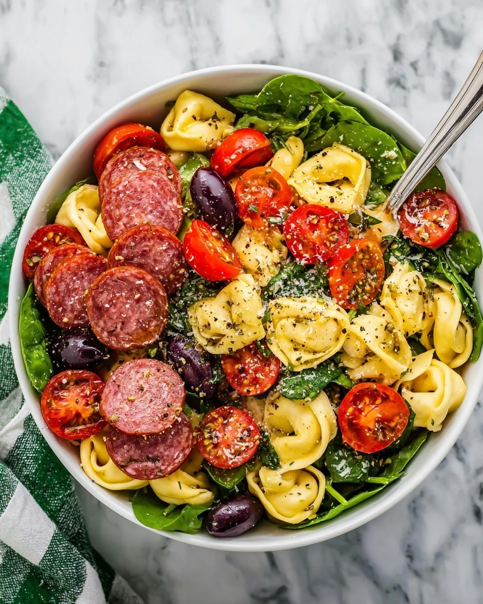 A white bowl filled with a colorful pasta salad shows three main layers: the base layer is fresh dark green spinach leaves, topped with a middle layer of creamy yellow tortellini pasta. The top layer has bright red cherry tomato halves, round slices of reddish salami with white specks, and dark black olives. The ingredients are evenly mixed and lightly sprinkled with ground black pepper and herbs. The bowl sits on a white marbled surface with a silver fork inside. photo taken with an iphone --ar 4:5 --v 7