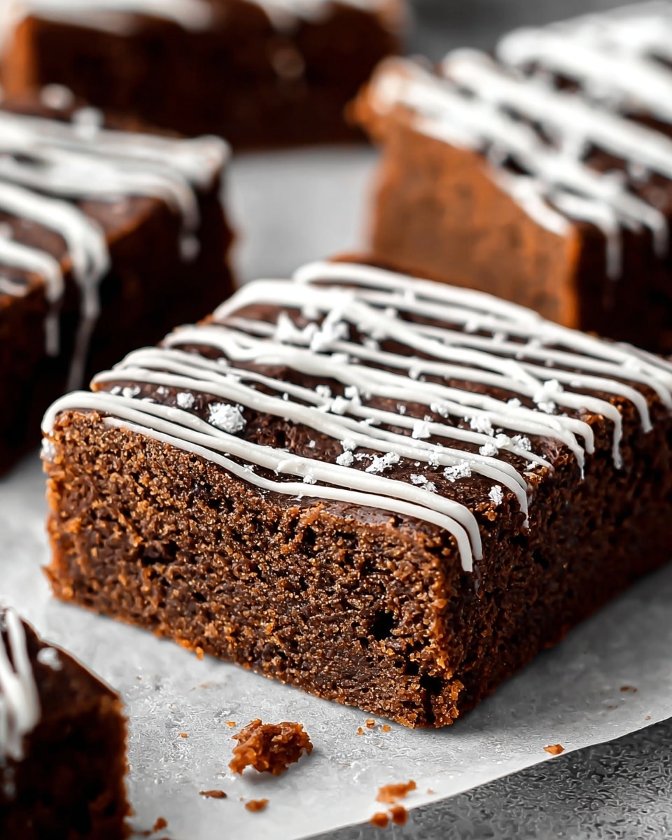 A close-up view of a thick brownie square with a rich, dark brown color and a slightly crumbly edge, sitting on a white marbled surface covered with a sheet of parchment paper. The top of the brownie is decorated with thin, zigzag lines of smooth white icing running from side to side. Small crumbs are scattered around the base of the brownie, showing its moist and dense texture inside. More similar brownie pieces are visible blurred in the background on the white marbled surface. photo taken with an iphone --ar 4:5 --v 7