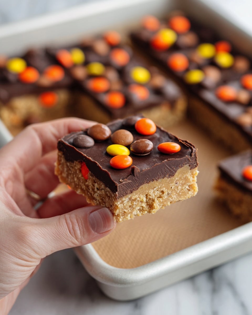 A close-up of a woman's hand holding a small square piece of a dessert bar with two layers: the bottom layer is thick and light brown with a crumbly texture, and the top layer is smooth dark chocolate with colorful round candy pieces in orange, yellow, and brown scattered on it. In the background, more of the dessert rests on a white baking tray lined with a brown silicone mat, all set on a white marbled surface. photo taken with an iphone --ar 4:5 --v 7