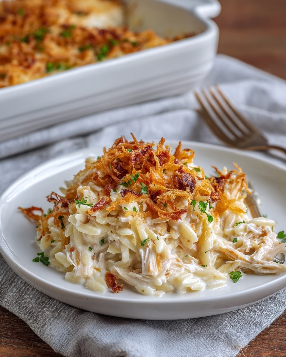 A white plate holds a creamy, cheesy dish made of orzo pasta mixed with shredded pieces of chicken in a thick white sauce. On top, there is a layer of crispy, golden-brown fried onions, lightly scattered and garnished with small bits of green herbs. The orzo looks soft and well-coated in the sauce, with some browned edges. The plate sits on a wooden surface with a light gray cloth partially underneath it. In the background, a white baking dish filled with the same food is slightly out of focus. Photo taken with an iphone --ar 4:5 --v 7