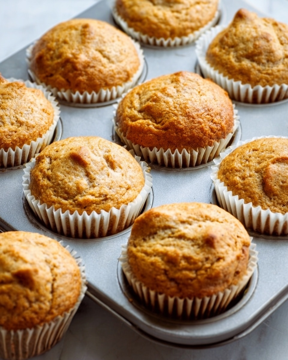 A close-up image of a baking tray filled with twelve golden brown muffins. Each muffin is nestled in white paper liners inside round metal cups. The muffins have a slightly cracked, domed top with a rough texture showing crumbs and small chunks inside. The muffins are packed closely together in a 3x4 grid on the baking tray. The tray sits on a white marbled surface. Photo taken with an iphone --ar 4:5 --v 7