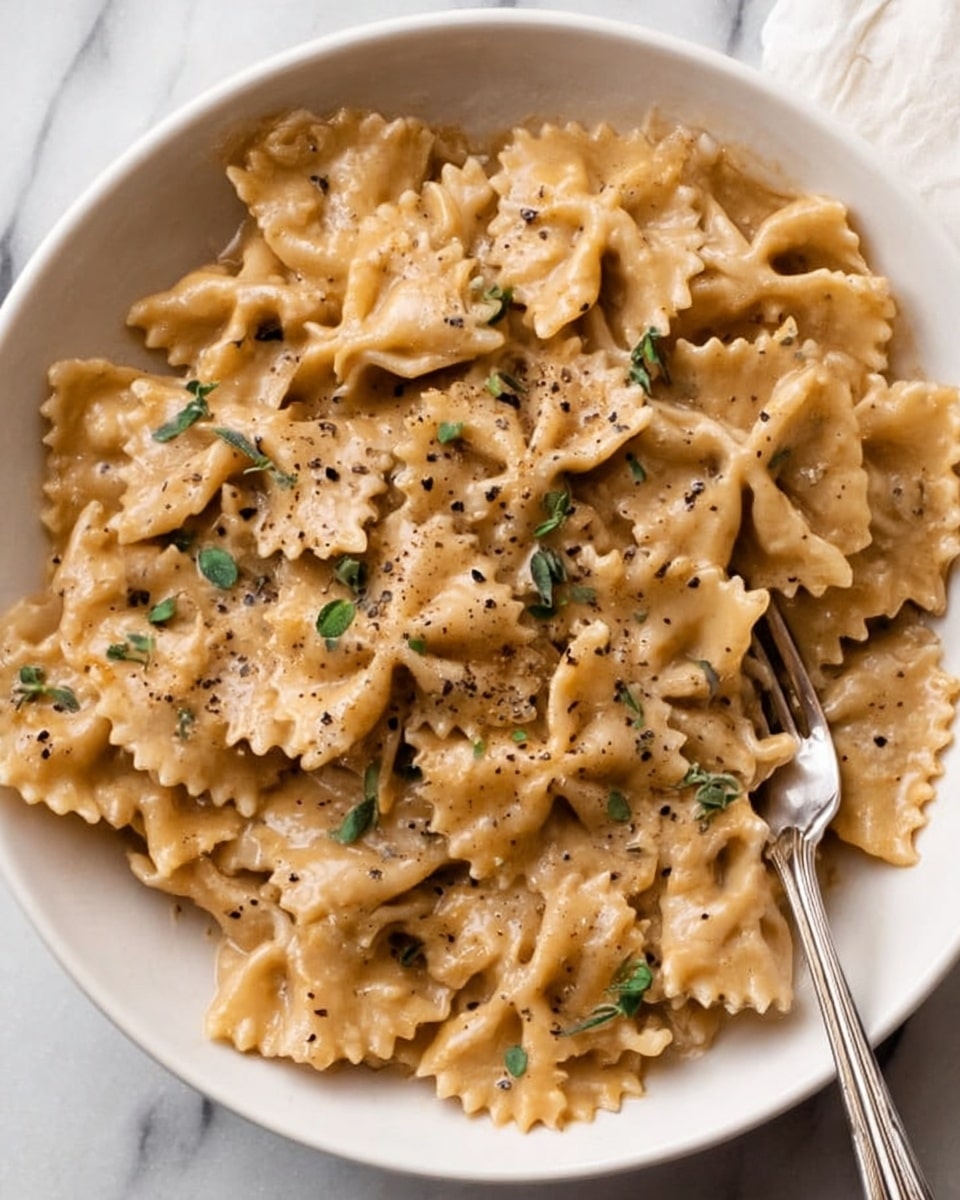 The dish is a white bowl filled with ruffled pasta pieces, covered in a creamy light brown sauce. The pasta layers have a slightly shiny texture from the sauce, with small green herb leaves scattered on top. There are many pasta pieces overlapping each other, and coarse black pepper is sprinkled over the entire dish. A silver fork rests on the right side inside the bowl, partially inside the pasta. The background is a white marbled surface. photo taken with an iphone --ar 4:5 --v 7