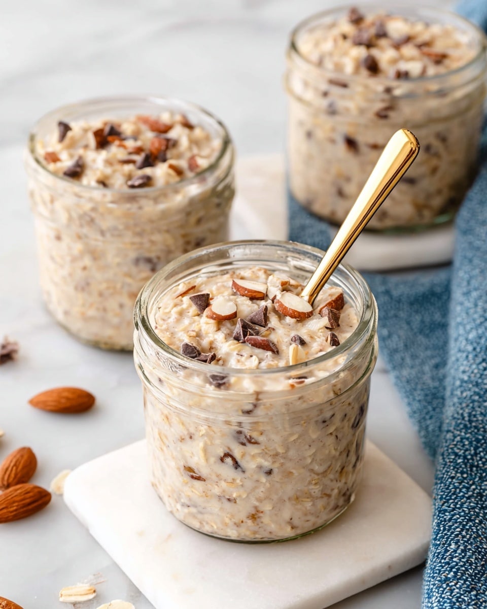 The image shows three clear glass jars filled with creamy overnight oats that have a light beige color mixed with small chunks of nuts and dark chocolate chips scattered throughout. The jar in the front is on a white square marble slab and holds oats mixed with visible almond pieces and tiny dark chocolate chips, with a gold spoon sticking out from the top right side. The texture looks thick and creamy with the oats and nuts evenly distributed. The background has a white marbled texture and there are a few loose chocolate chips and an almond on the surface near the jars. photo taken with an iphone --ar 4:5 --v 7