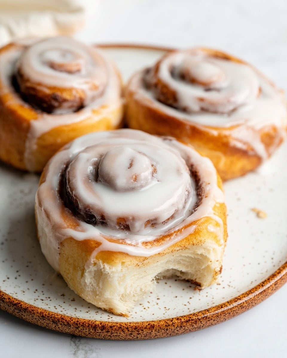 Three cinnamon rolls sit on a white plate with a speckled brown rim, placed on a white marbled surface. Each roll has a thick spiral shape with visible layers of golden-brown dough and darker cinnamon filling. The rolls are generously coated with glossy white icing that drips slightly down the sides, adding a smooth and creamy texture on top. The front roll has a bite taken out, showing the soft, fluffy inside with a moist crumb. The background is softly blurred, highlighting the warm, inviting look of the pastries. photo taken with an iphone --ar 4:5 --v 7