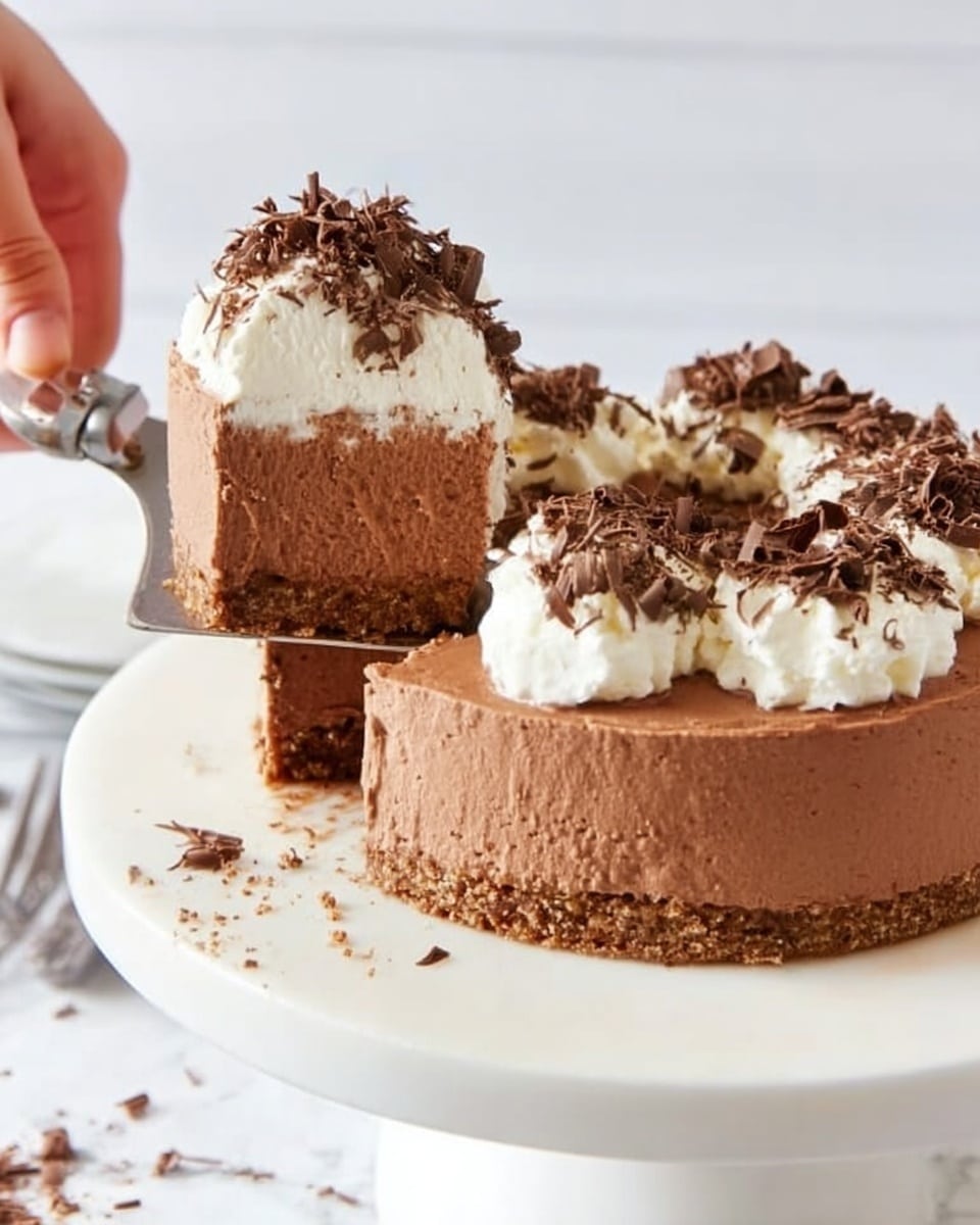 A thick slice of creamy chocolate mousse cake is being lifted by a silver cake server over a round white plate holding the rest of the cake. The cake has three layers: the bottom is a thin, light brown crumbly crust; the middle is a thick, smooth milk chocolate mousse layer with small air bubbles and a few cracks; the top is a fluffy white whipped cream dollop with dark chocolate shavings sprinkled on it. More chocolate shavings are scattered around the plate on a white marbled surface. The background is plain white wood panels. photo taken with an iphone --ar 4:5 --v 7