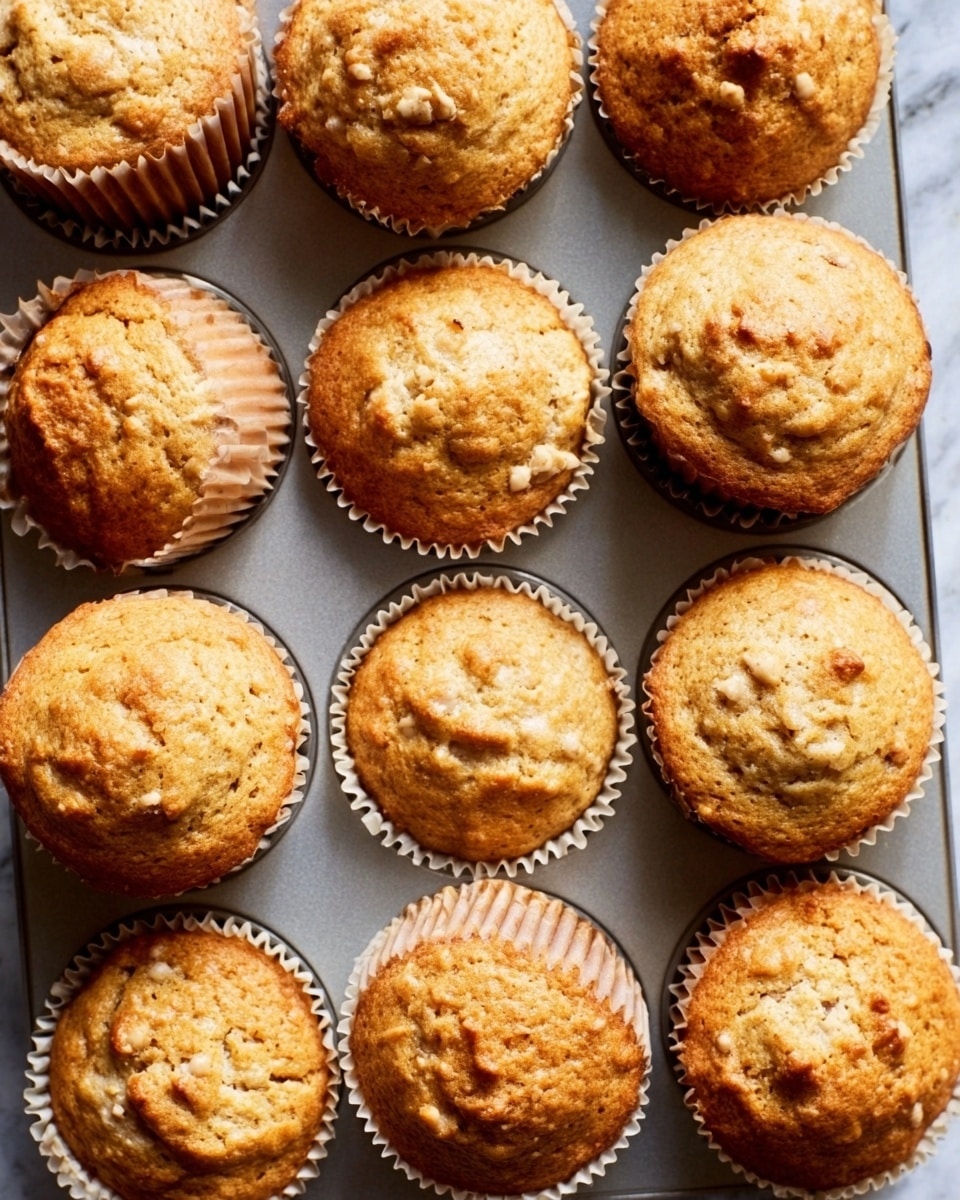 A silver muffin tray holds twelve golden brown muffins with slightly cracked tops showing a soft, crumbly texture. Each muffin is seated in white paper liners, and the muffins look freshly baked with some slightly tilted to one side, revealing their soft inner crumb. The tray is set on a white marbled surface, giving a clean and bright background that highlights the warm tones of the muffins. photo taken with an iphone --ar 4:5 --v 7