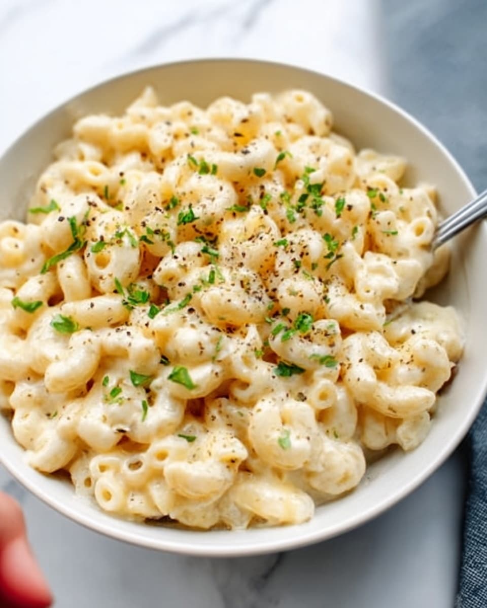 The image shows a close-up of a bowl filled with creamy macaroni and cheese. The dish has short, curved pasta in a thick, smooth, light yellow cheese sauce that looks rich and creamy. The top layer is sprinkled with finely chopped green herbs and a little black pepper, adding small green and black dots all over the pasta. The bowl is white and round, sitting on a white marbled surface, with part of a woman's hand holding a spoon just visible, ready to scoop some of the macaroni. Photo taken with an iphone --ar 4:5 --v 7