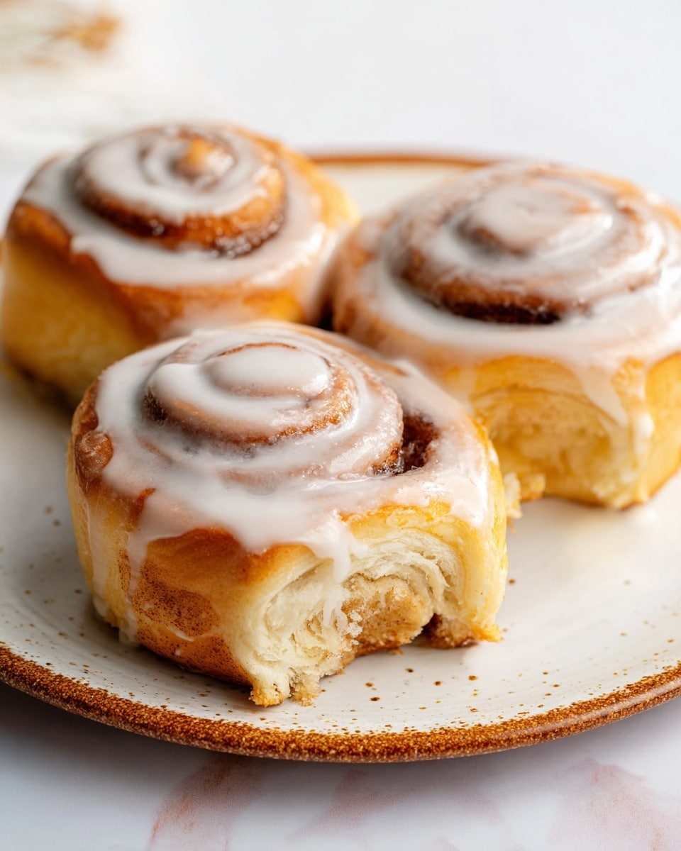 A close-up view of three cinnamon rolls on a white plate, each roll showing a thick spiral shape with a golden-brown base layer of dough that looks soft and fluffy, topped with a light brown cinnamon layer arranged in a swirl pattern, and covered with a glossy white icing layer that drips slightly down the sides, making the rolls look moist and sweet; the background features a clean white marbled texture. photo taken with an iphone --ar 4:5 --v 7