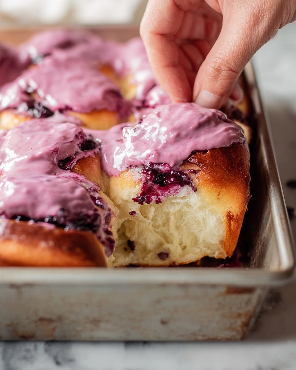 A close-up image shows a woman's hand pulling apart a soft, light golden brown bun with a fluffy and airy inside texture, topped with a thick layer of creamy purple-pink frosting that has a smooth but slightly lumpy texture. The bun sits in a gray metal baking tray with more buns and frosting visible in the background. Some dark purple berry filling is seen oozing out from under the frosting near the bottom of the bun. The tray rests on a white marbled surface. photo taken with an iphone --ar 4:5 --v 7