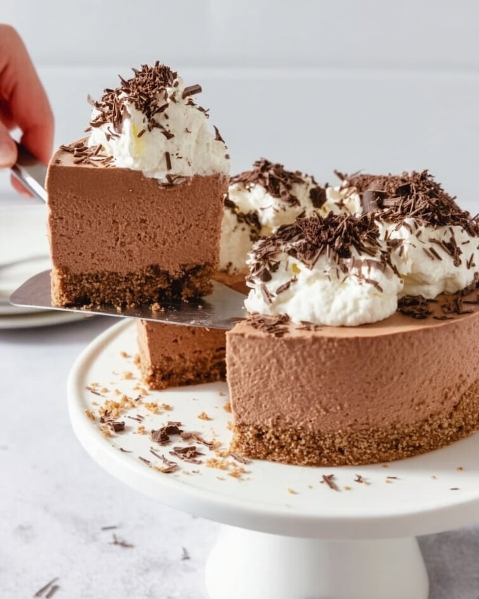 A rich chocolate mousse cake is shown on a white round cake stand placed on a white marbled surface. The cake has three visible layers: a thin crumbly light brown crust at the bottom, a thick smooth medium brown chocolate mousse filling in the middle, and fluffy white whipped cream dollops on top. The whipped cream is decorated with dark chocolate shavings sprinkled generously over it, with extra shavings scattered on the cake stand around the cake. A slice of the cake is being lifted with a silver cake server, revealing the clean, creamy texture of the chocolate mousse and the crumbly crust. A woman's hand is holding the cake server from the left side. The background is light and simple to focus attention on the cake. photo taken with an iphone --ar 4:5 --v 7